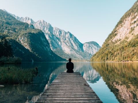 Von hinten sichtbare Person am Ende eines Holzstegs auf einem ruhigen See mit Spiegelung der Berge.